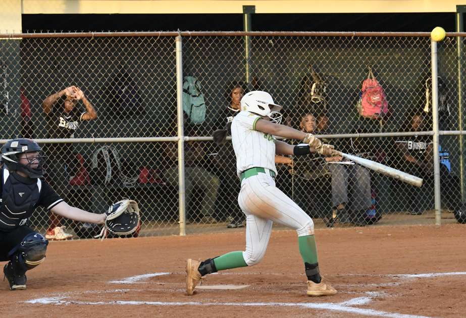 A softball player in white uniform swings at a pitch, with a catcher and spectators visible in the background behind a chain-link fence.