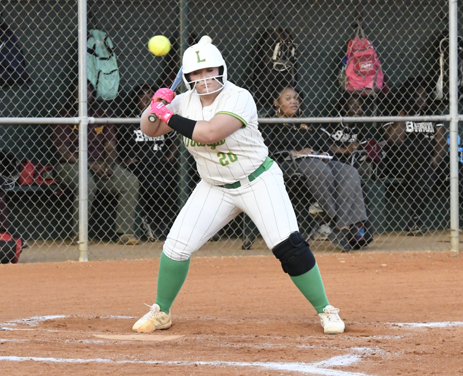 Softball player in white and green uniform prepares to bunt a pitch during a game, with teammates and equipment in the dugout behind a chain-link fence.