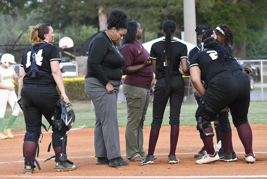 A softball team huddles on the field with coaches, discussing strategy during a game. Players wear black uniforms and catcher's gear. Trees and other players are visible in the background.