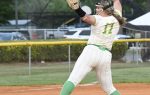 A softball pitcher in a white and green uniform winds up to throw a pitch on a dirt field during a game.