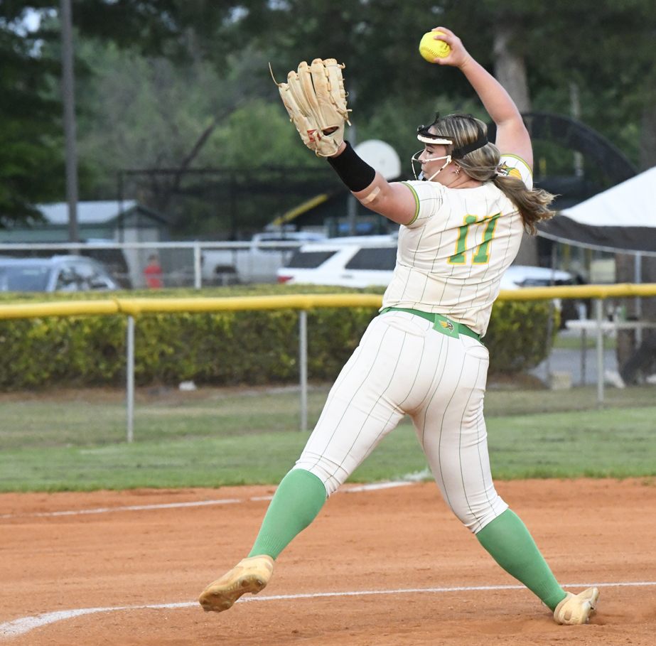 A softball pitcher in a white and green uniform winds up to throw a pitch on a dirt field during a game.