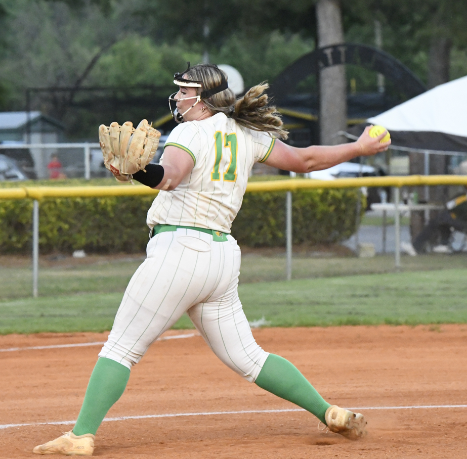 A softball player in a white and green uniform winds up to pitch on a dirt field, wearing a faceguard and glove.