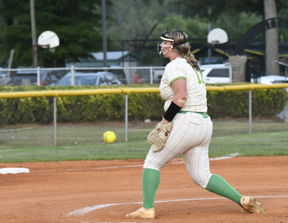 A softball pitcher in a white and green uniform throws a pitch on a dirt field, with yellow softball mid-air. Basketball hoops are visible in the background.