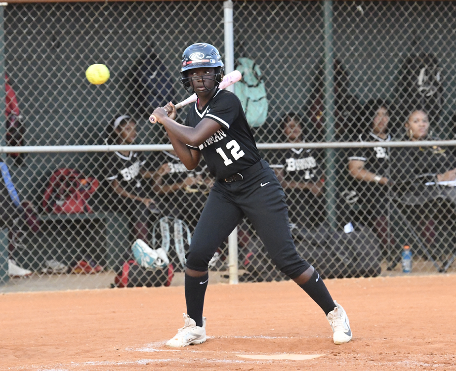 A softball player wearing number 12 prepares to bat as a yellow softball approaches, with teammates watching from the dugout behind a chain-link fence.