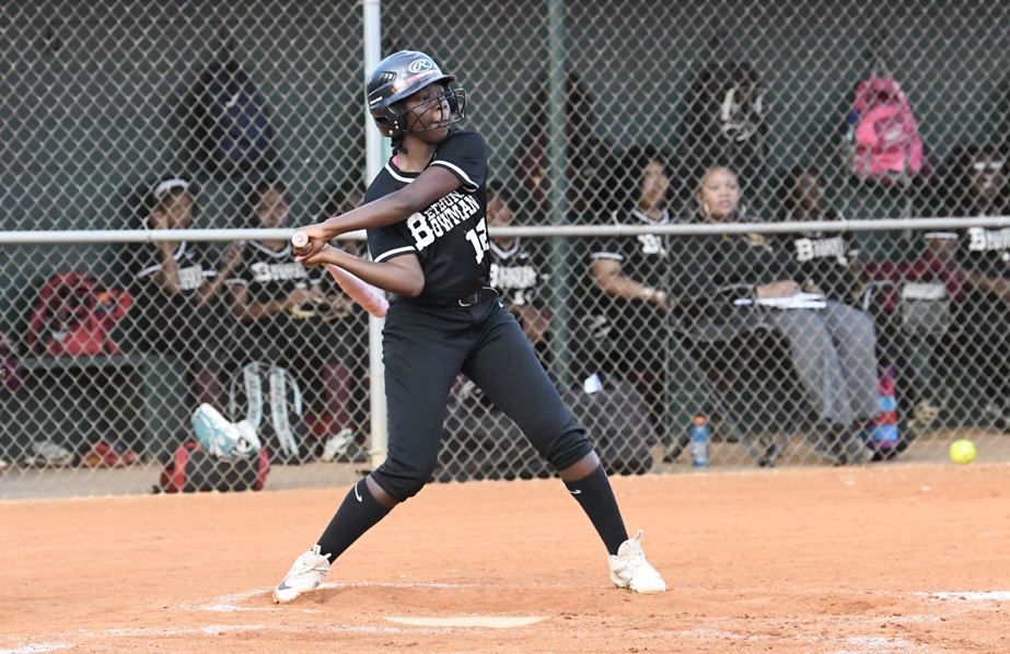 A softball player in a black uniform swings a bat during a game, with teammates watching from the dugout behind a chain-link fence.