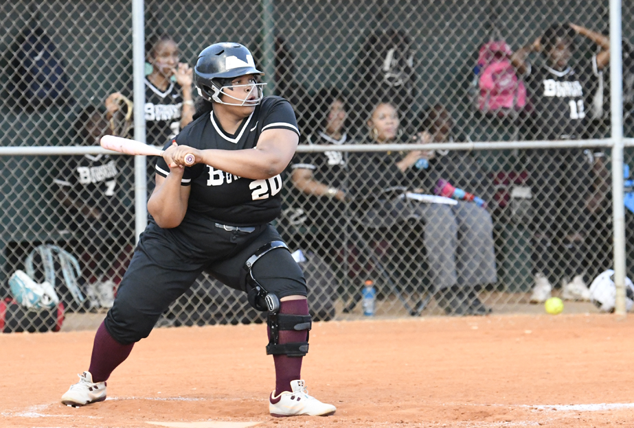 A softball player in black uniform prepares to bat, with teammates sitting behind a chain-link fence in the dugout.