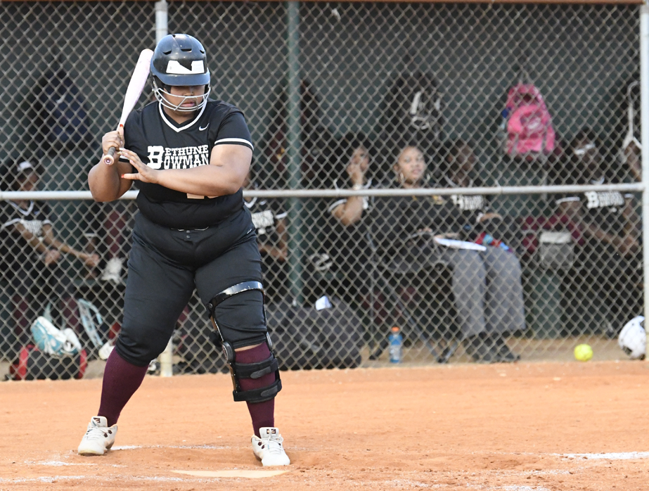A softball player in black uniform prepares to bat, wearing a helmet and knee brace, with teammates watching from the dugout behind a chain-link fence.