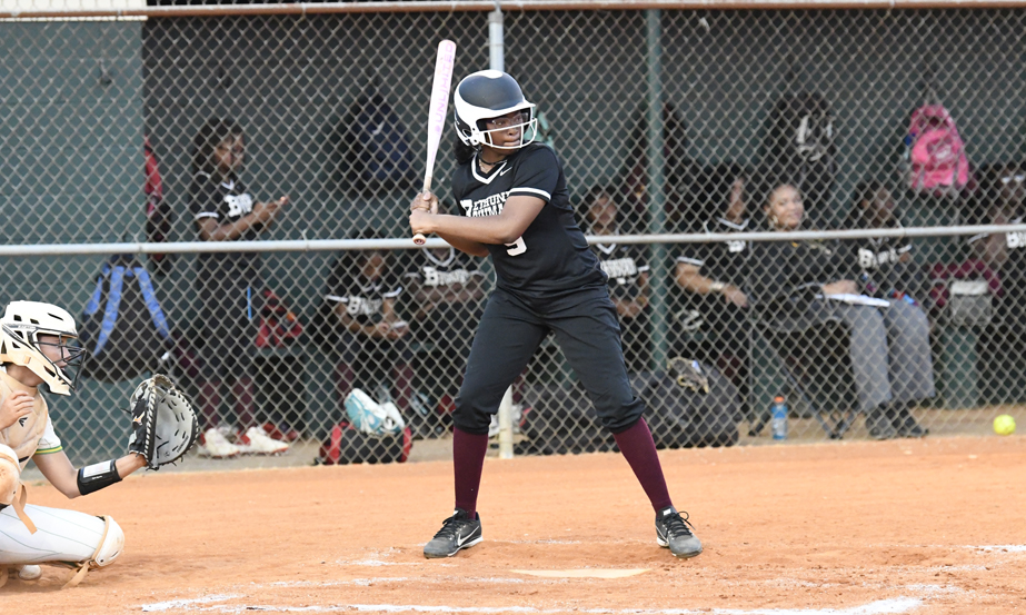 A softball player in black uniform stands ready to bat while a catcher and team members watch from the dugout behind a chain-link fence.
