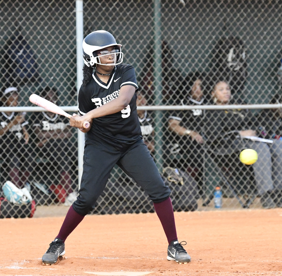 A softball player in black uniform prepares to hit a pitched yellow ball during a game, with teammates watching from the dugout behind a chain-link fence.