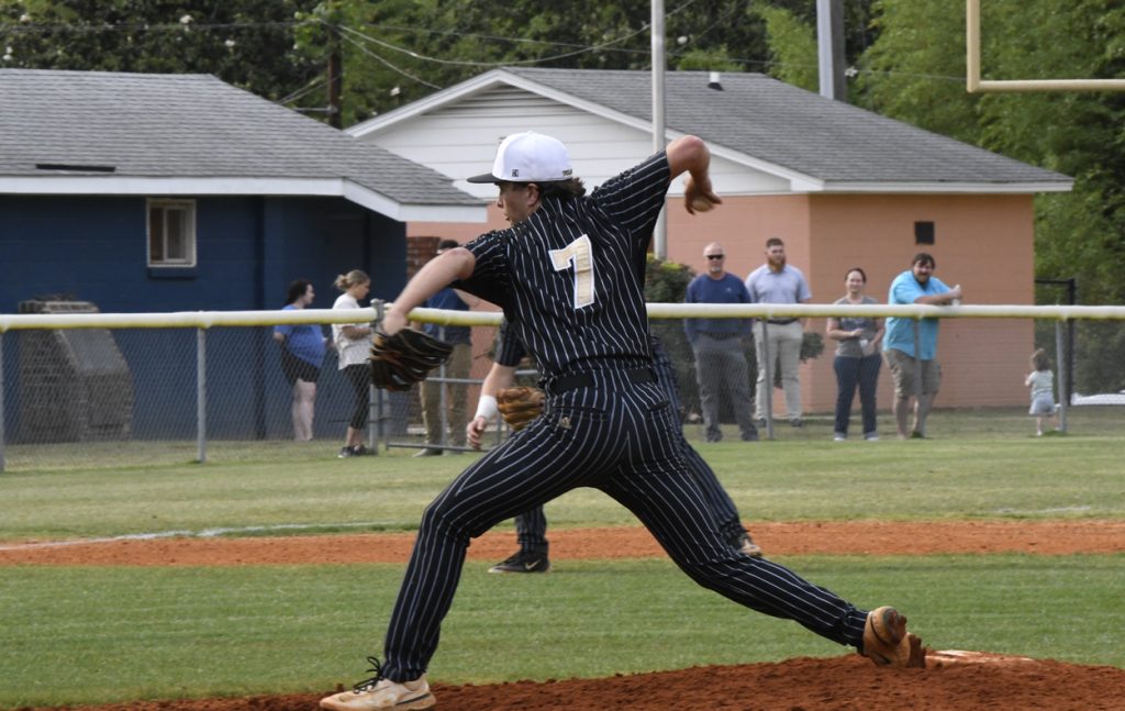 Baseball pitcher in a striped uniform throwing a pitch on a field, with spectators and buildings in the background.