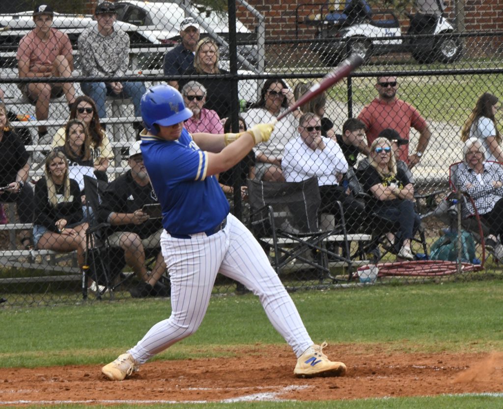 Baseball player in blue jersey swings bat during a game, with spectators watching from the stands behind a chain link fence.