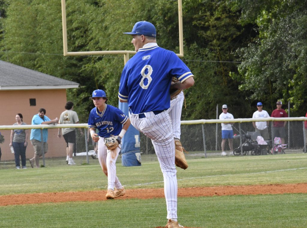 Baseball pitcher in blue uniform prepares to throw, while another player and spectators watch on a sunny outdoor field.