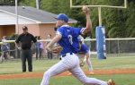 Baseball pitcher in blue uniform throws a pitch on a field, with umpire, teammates, and spectators in the background.