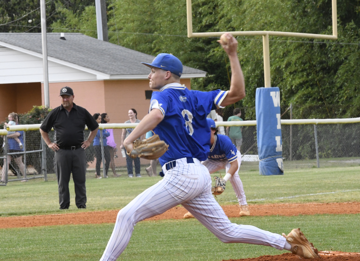 Baseball pitcher in blue uniform throws a pitch on a field, with umpire, teammates, and spectators in the background.