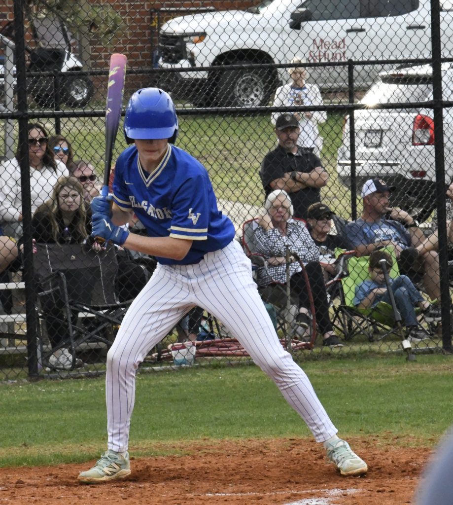 Baseball player in blue uniform preparing to bat, with fans seated and standing behind a fence, and parked cars visible in the background.