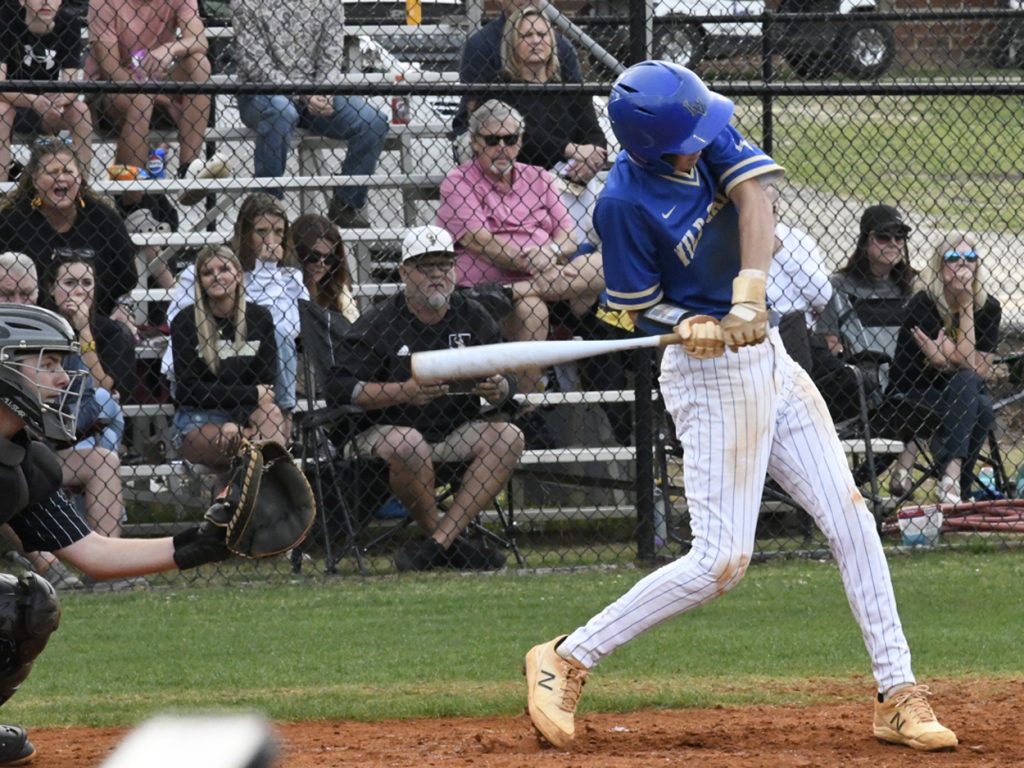 Baseball player in blue uniform swings bat at pitch, while catcher and crowd watch from bleachers behind a chain-link fence on a sunny day.