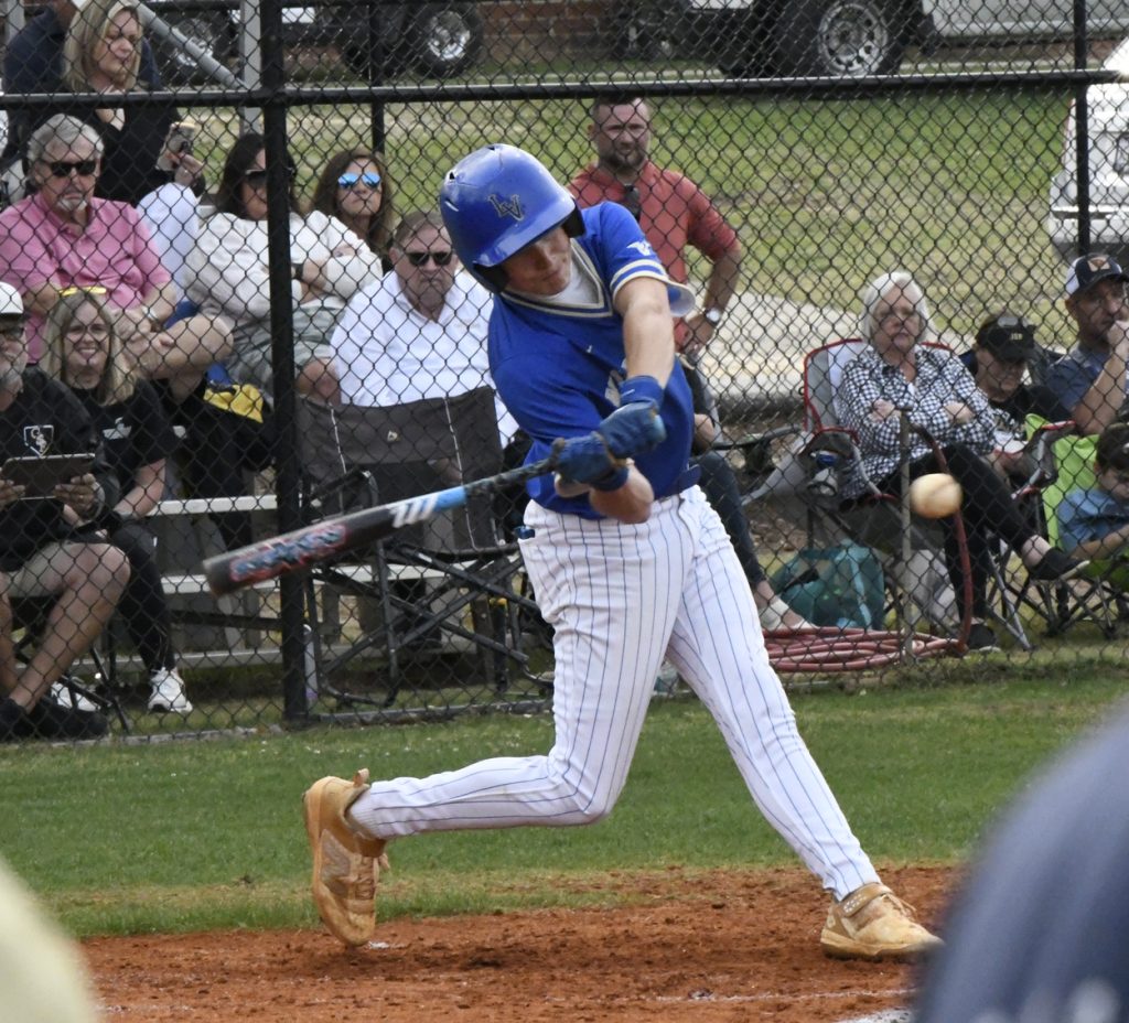 Baseball player in blue uniform swinging bat at a pitched ball during a game, with spectators watching from behind a fence in the background.