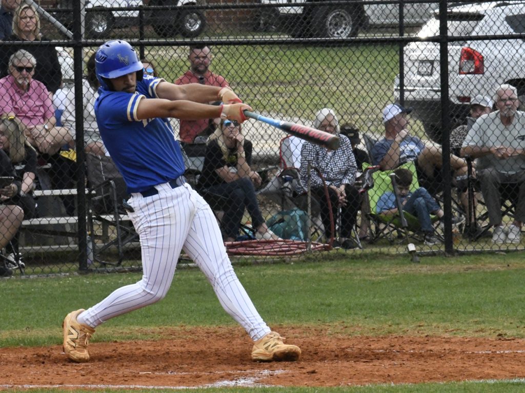 A baseball player in blue uniform swings a bat during a game, with spectators watching behind a chain-link fence.