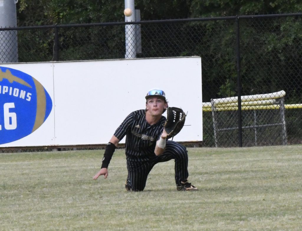 A baseball player in black pinstripe uniform crouches in the outfield, focused and ready to catch a fly ball with glove raised.