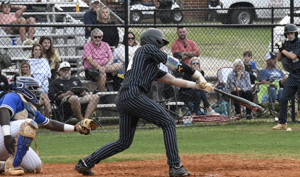 Baseball player in black pinstripes swinging bat, catcher in blue gear, spectators seated in bleachers behind a chain-link fence watching the game.