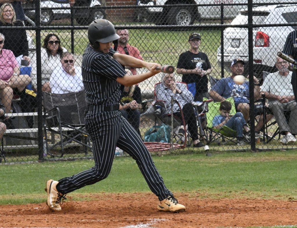 A baseball player in a black pinstripe uniform swings at a pitch, with spectators watching behind a fence.