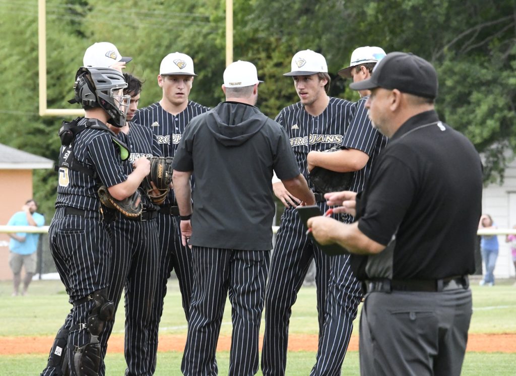Baseball team in black pinstripe uniforms huddles with coach on field; umpire stands nearby holding a notepad; trees and goalposts in background.