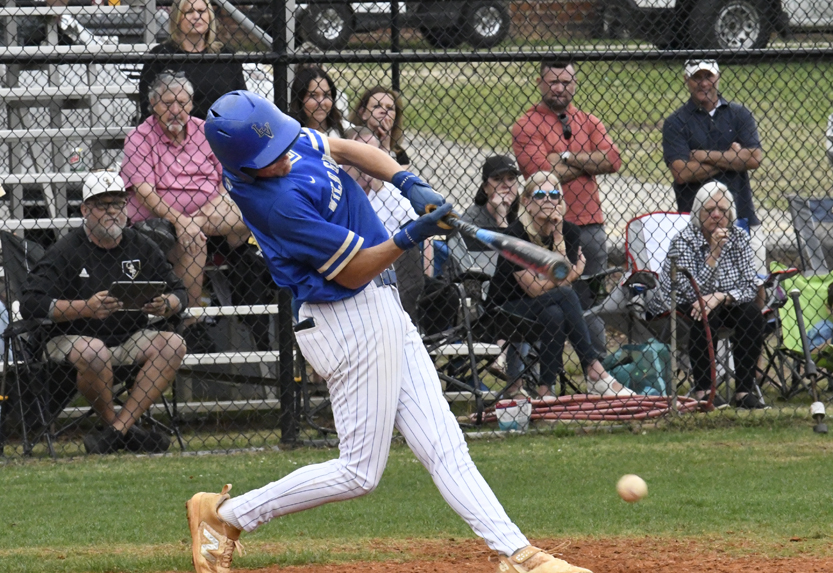 A baseball player in blue uniform swings at a pitch during a game, with spectators watching from the stands and behind a chain-link fence.