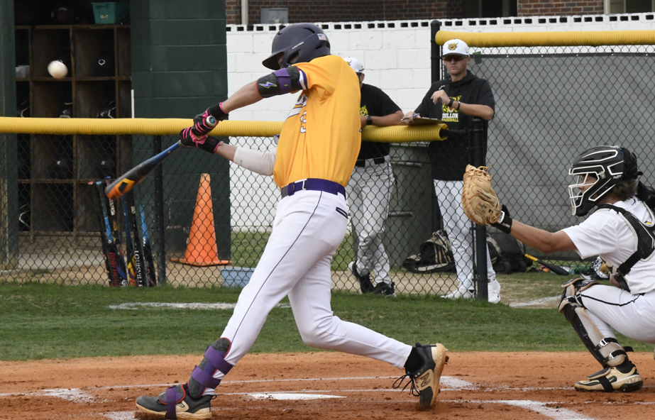 Baseball player in yellow jersey swings bat at pitch, with catcher and onlookers in background during a game.
