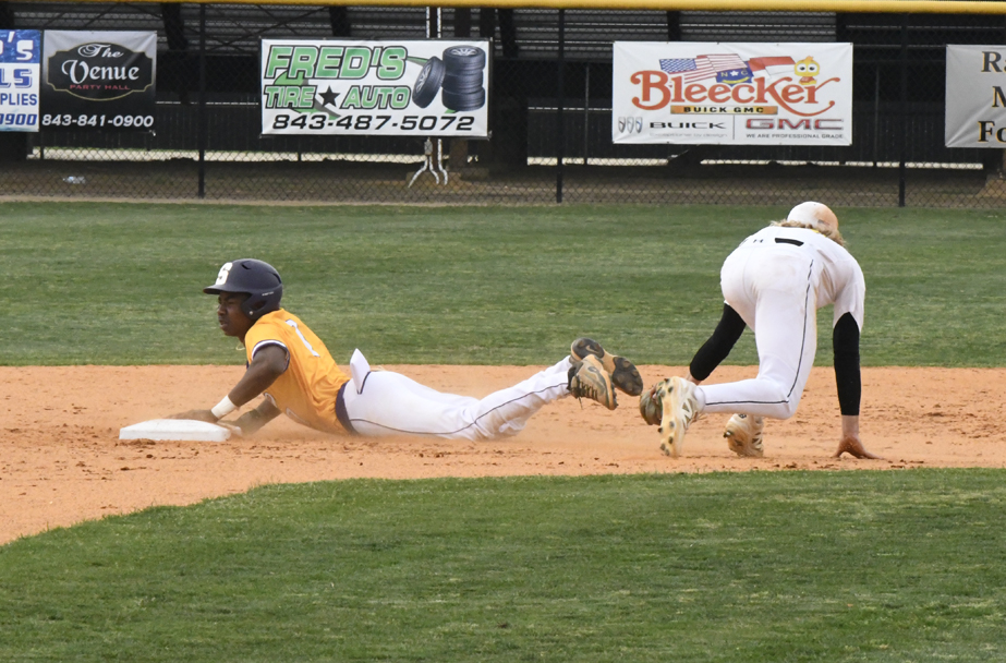 A baseball player in a yellow jersey slides safely into a base as a fielder in white bends down to catch the ball.