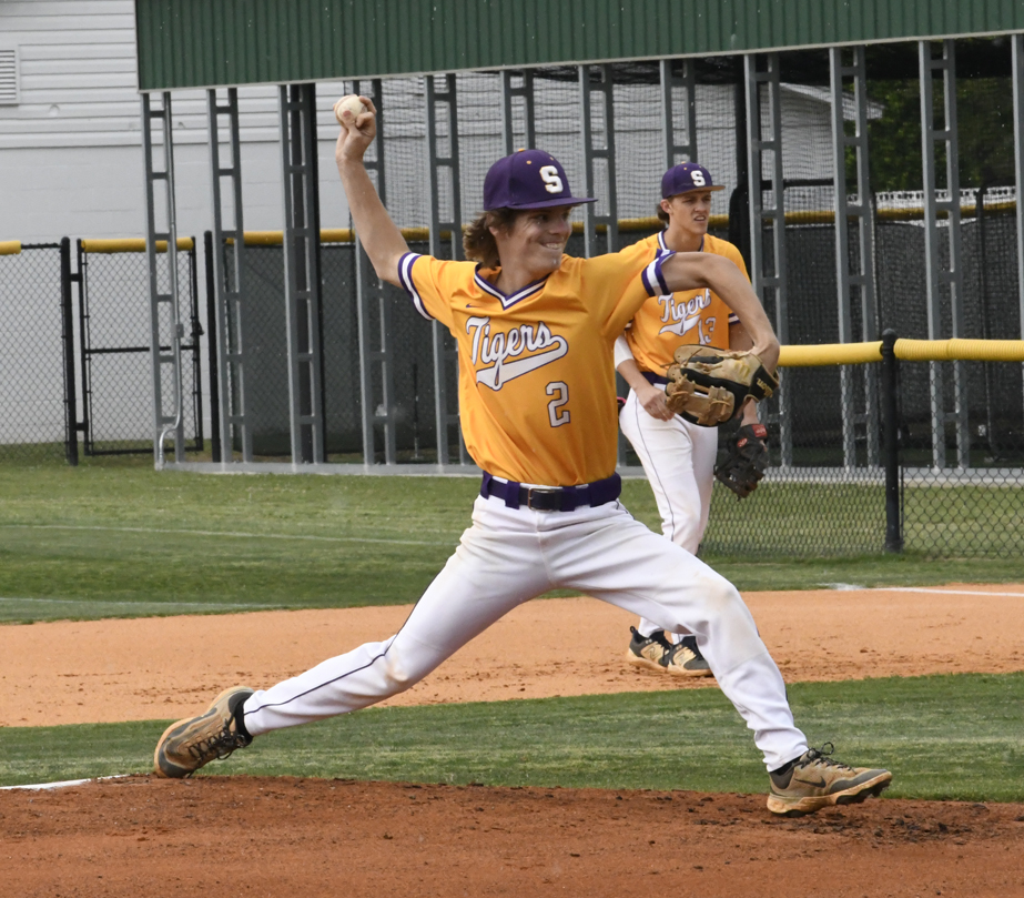 A baseball pitcher in a yellow "Tigers" jersey winds up to throw, with a teammate in the background on the field.