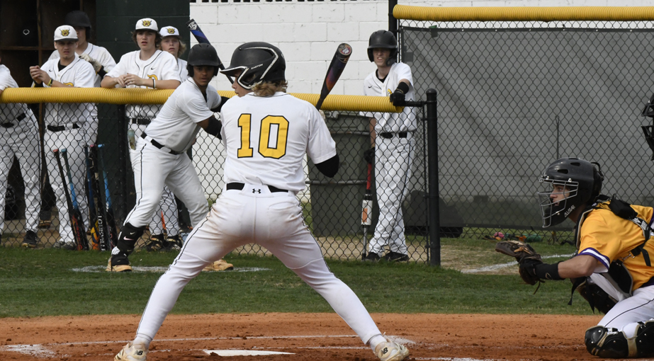 A baseball player at bat in a game, facing a catcher and pitcher, with teammates watching from the dugout in the background.