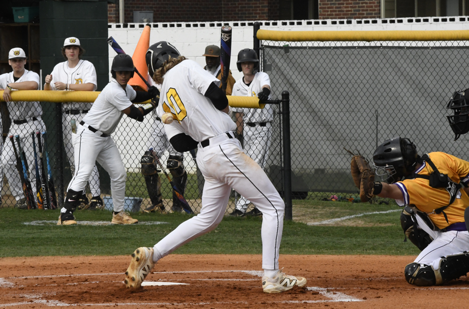 A baseball batter in white uniform swings at a pitch while the catcher and other players watch from behind the fence.