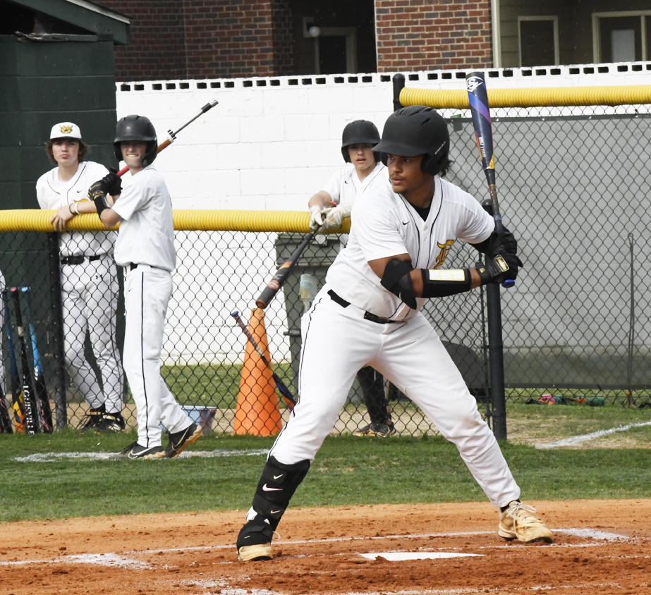 A baseball player in white uniform prepares to bat, while teammates watch and practice behind a chain-link fence on a sunny day.