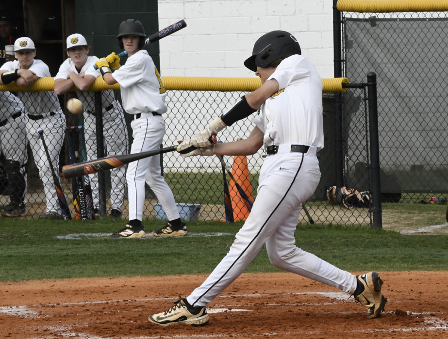 A young baseball player in a white uniform swings at a pitch during a game, while teammates watch from the dugout in the background.