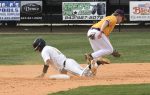 A baseball player in white slides safely onto a base as a player in yellow jumps to avoid him during a game.