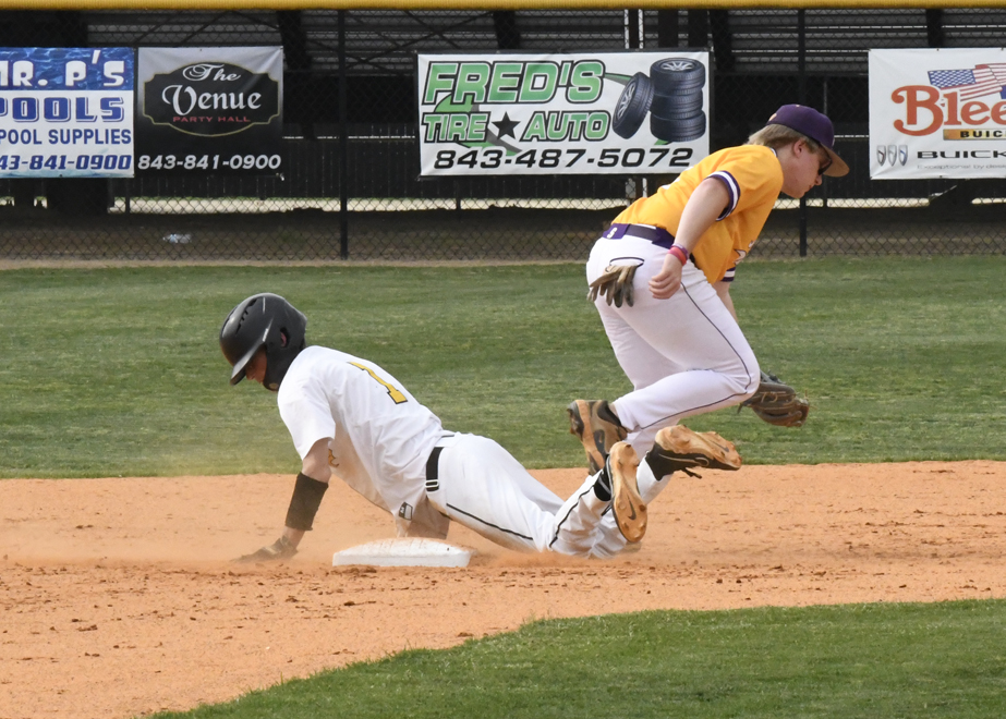 A baseball player in white slides safely onto a base as a player in yellow jumps to avoid him during a game.