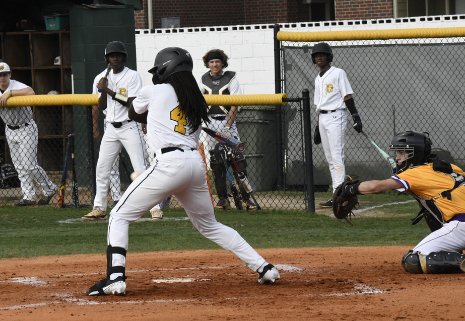 A baseball player swings at a pitch during a game while the catcher and several teammates watch from behind the fence.