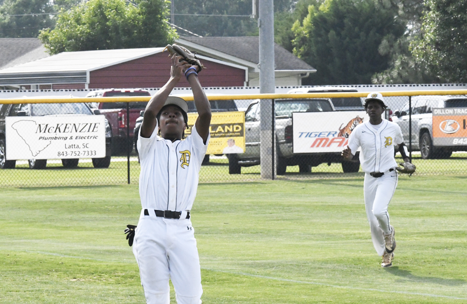 Two young baseball players in white uniforms during a game, with one catching a ball and the other running toward him on a grassy field.