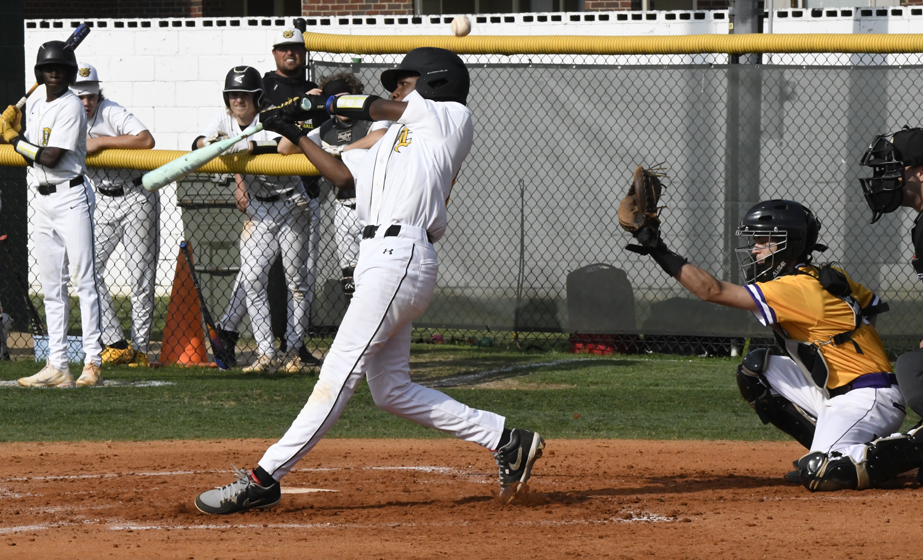 A baseball batter in white swings at a pitch, with a catcher and umpire positioned behind home plate and teammates watching from the dugout.