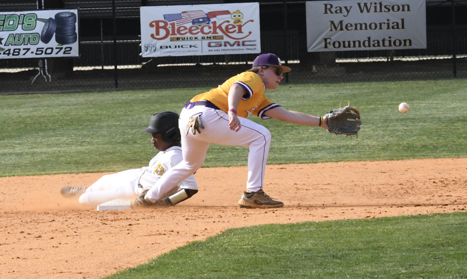 A baseball player in yellow attempts to tag a sliding runner in white as the ball approaches on a dirt infield.