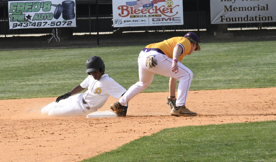 Baseball player in white uniform slides safely into a base while another player in yellow attempts to tag him out on a dirt infield.