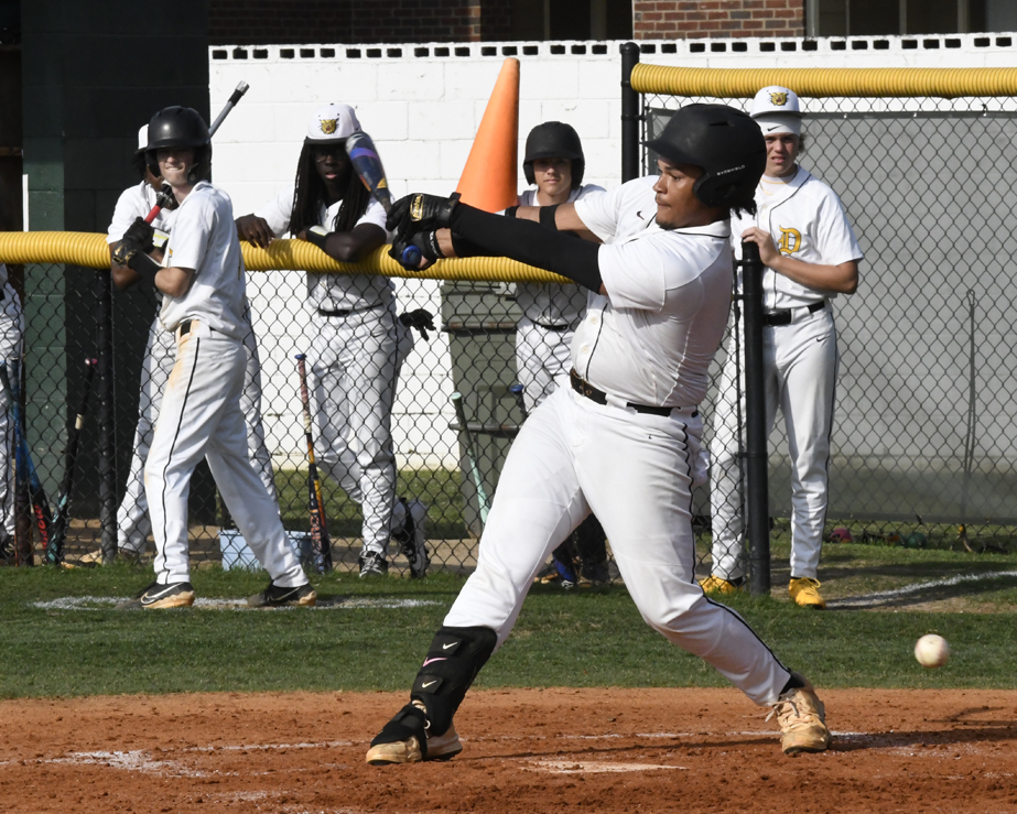 Baseball player in white uniform swings at a pitch while teammates watch from behind the fence on a sunny day.