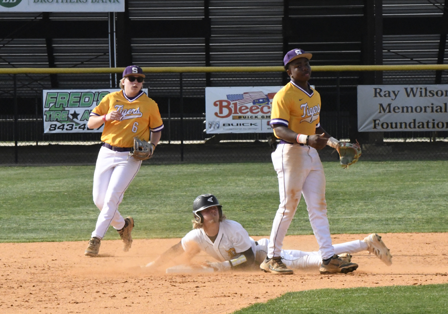 A baseball player slides into base as two opposing players in yellow uniforms watch and prepare to make a play on a sunny field.