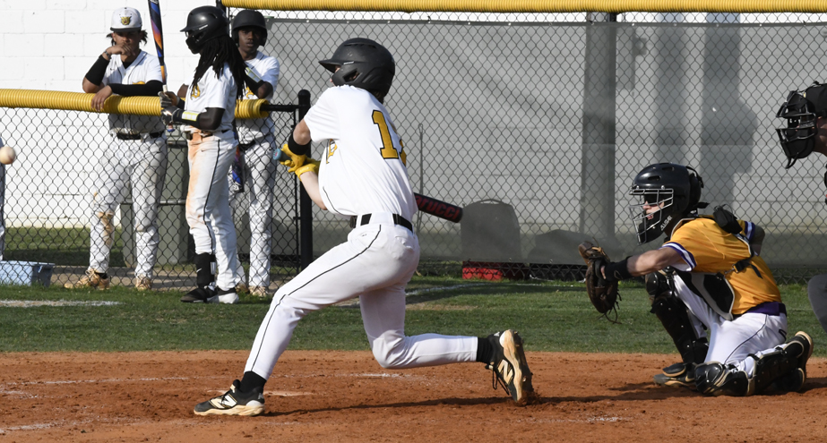 A baseball player in a white uniform swings at a pitch, while the catcher and umpire kneel behind home plate. Teammates watch from behind a fence.