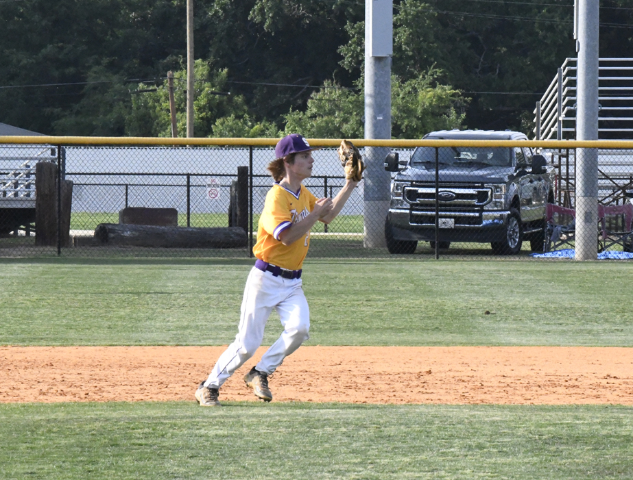 Baseball player in yellow and white uniform preparing to catch a ball on a field, with a fence and parked truck in the background.