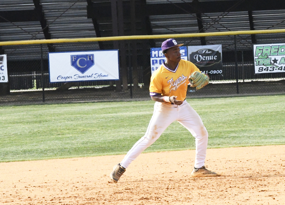 Baseball player in a yellow "Tigers" jersey and white pants prepares to throw a ball on a field, with empty bleachers and signs in the background.