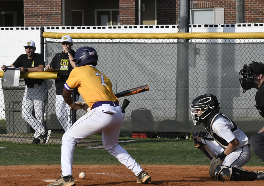 Baseball player in yellow jersey bunting at home plate, catcher and umpire ready behind, teammates watching from dugout in background.