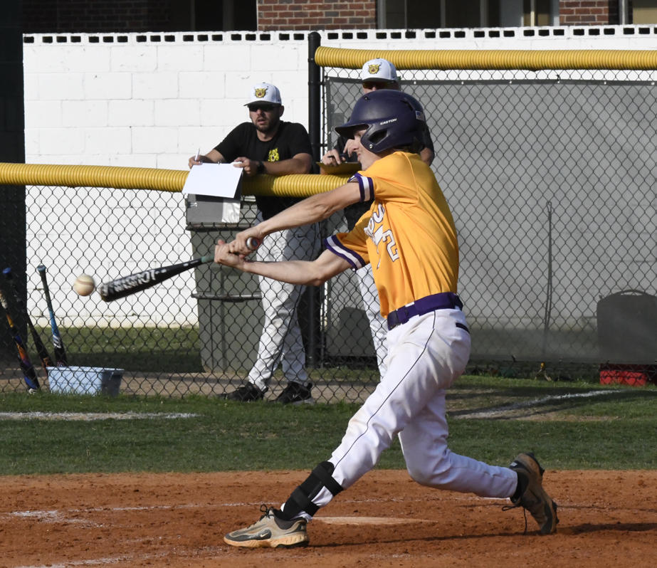 Baseball player in a yellow jersey swings and hits the ball during a game, with coaches watching behind a chain-link fence.