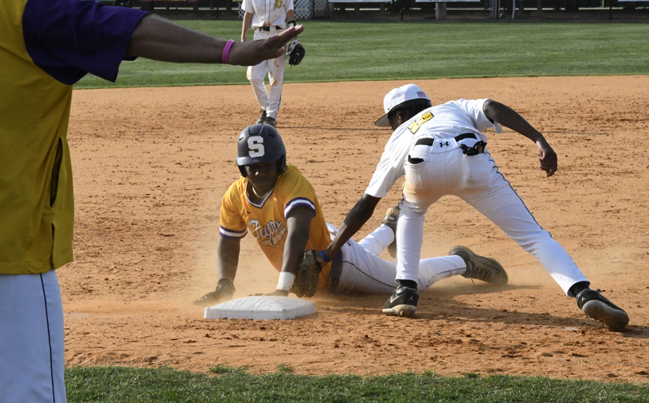 A baseball player in yellow slides into a base as a player in white attempts to tag him, with dust rising and an umpire's arm signaling in the foreground.
