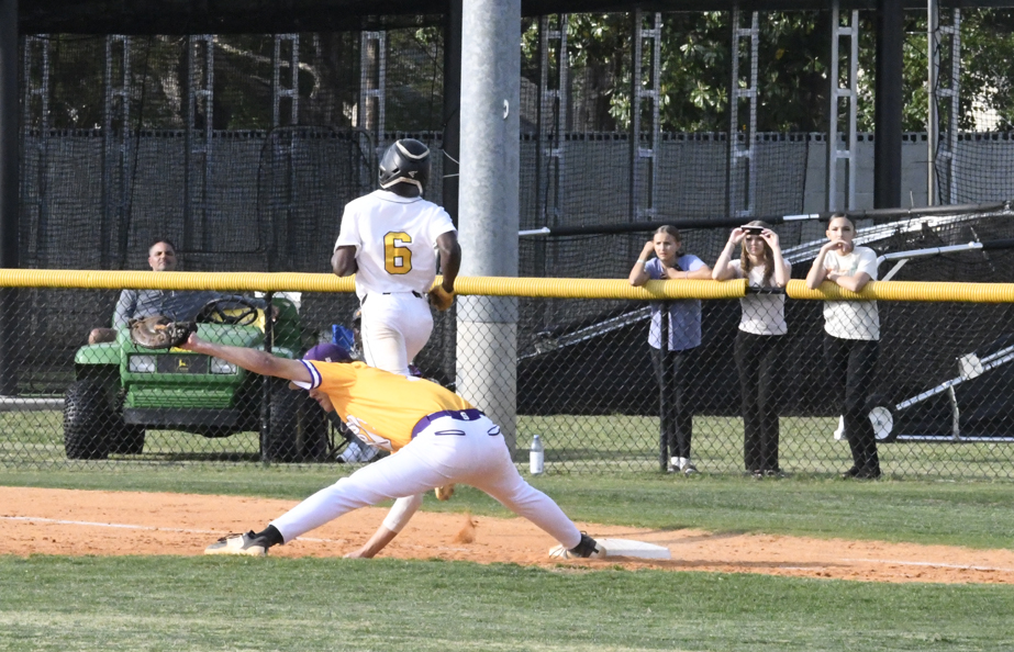 Baseball player in white runs to base as player in yellow stretches to catch ball; spectators watch behind fence.